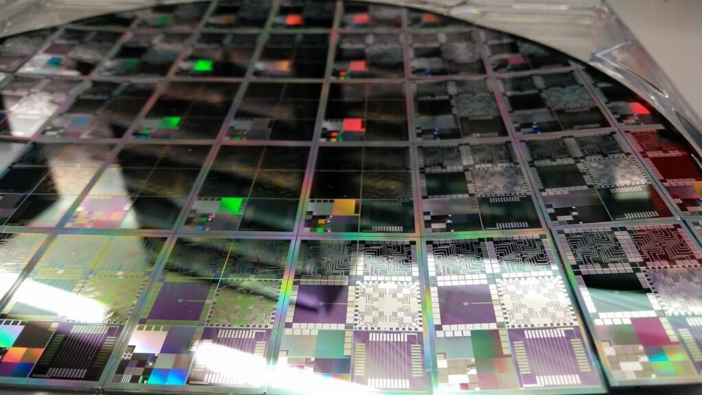 A close-up photograph of a large circular 12-inch semiconductor wafer containing multiple microelectronic testbed die, displaying iridescent multicoloured reflections and intricate circuit patterns across its surface, held in a clear plastic carrier