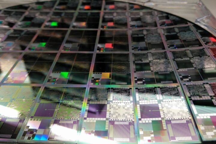 A close-up photograph of a large circular 12-inch semiconductor wafer containing multiple microelectronic testbed die, displaying iridescent multicoloured reflections and intricate circuit patterns across its surface, held in a clear plastic carrier