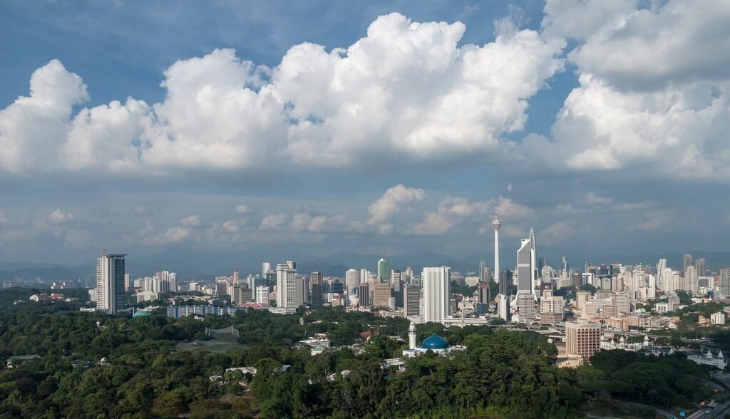 Aerial panoramic view of the Kuala Lumpur city skyline, Malaysia, featuring high-rise buildings, the KL Tower, lush green trees in the foreground, a mosque with a blue dome, and a dramatic cloudy blue sky above