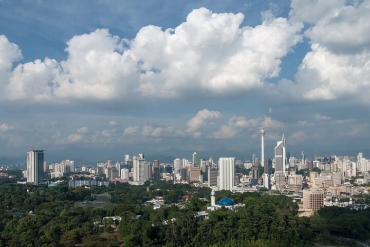 Aerial panoramic view of the Kuala Lumpur city skyline, Malaysia, featuring high-rise buildings, the KL Tower, lush green trees in the foreground, a mosque with a blue dome, and a dramatic cloudy blue sky above